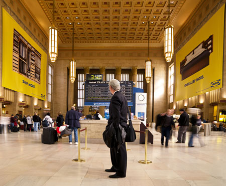 A man in a dark suit standing still in a busy station hall under the departures board